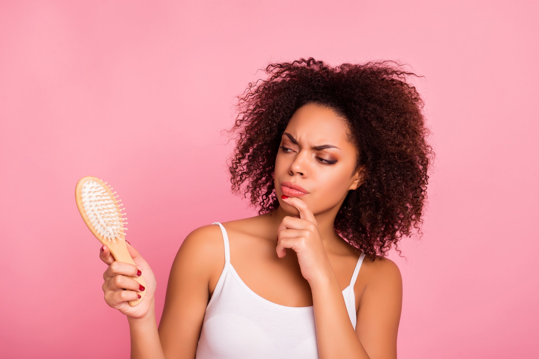 Attractive, pretty, charming, thoughtful, unhappy, sad girl looking at comb in hand touching chin with finger, having dry, oiled hair loss, she need mask, lotion, balm, isolated on pink background